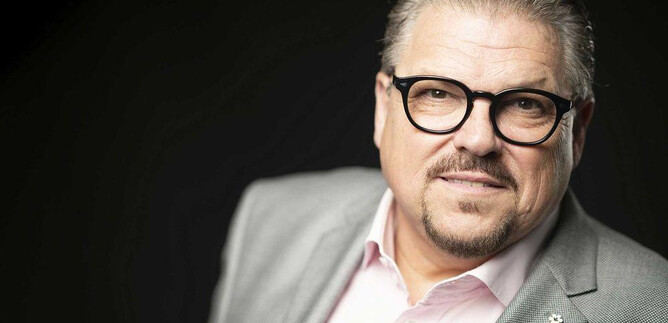 A man with glasses and a well-groomed appearance smiles slightly. He is wearing a gray suit and a light-colored shirt. | © Lukas Beck