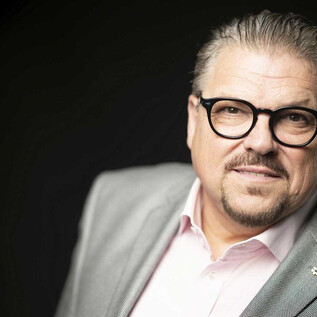 Muž s brýlemi a upraveným vzhledem se mírně usmívá. Nosí šedý oblek a světlou košili. | © Lukas Beck