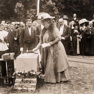 Kobieta w eleganckim stroju kładzie kamień na stole, podczas gdy duża grupa ludzi w tle obserwuje. Scena najwyraźniej rozgrywa się podczas uroczystego wydarzenia lub ceremonii. | © ISM