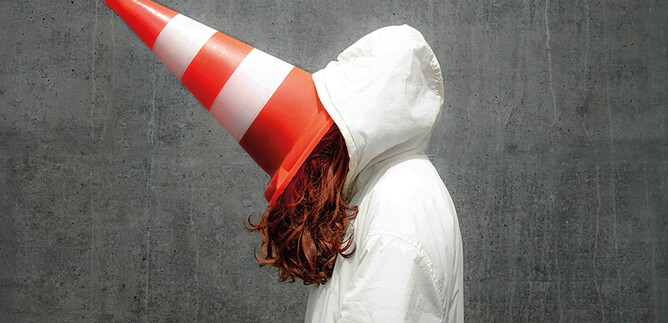 Egy személy fehér kabátot visel, és közúti forgalmi kúp van a fején. A háttér szürke és egyszerű. | © Valentina Schuster