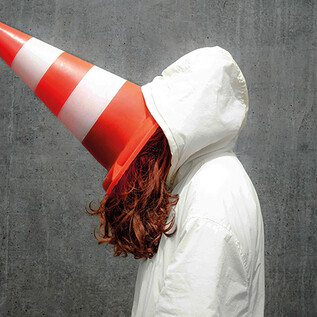 A person is wearing a white jacket and has a traffic cone on their head. The background is gray and plain. | © Valentina Schuster