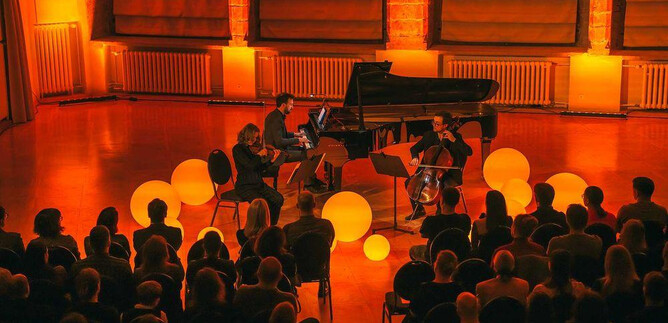 A live music performance featuring three musicians on stage. The space is surrounded by warm light and decorative balls, while the audience follows the performance. | © Freie Kunst