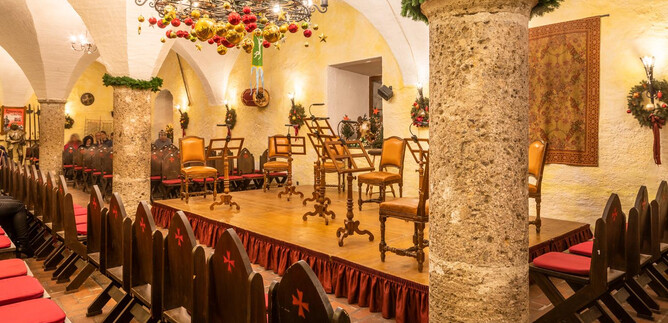 A beautiful hall with wooden chairs and a festive chandelier. The walls are decorated and create a welcoming atmosphere. | © salzburghighlights