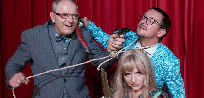 Three people in a funny scenario, with one holding a weapon and the other tied up with a rope. In the background, a red curtain can be seen. | © Sigrid Riepl