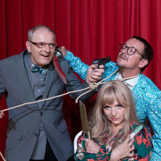 Three people in a funny scenario, with one holding a weapon and the other tied up with a rope. In the background, a red curtain can be seen. | © Sigrid Riepl