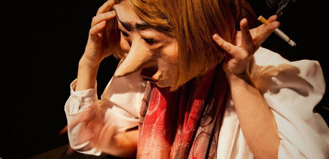 A person with a large mask is sitting in front of a laptop. They look stressed and are holding a cigarette in their hand. | © Debra Tchotchov