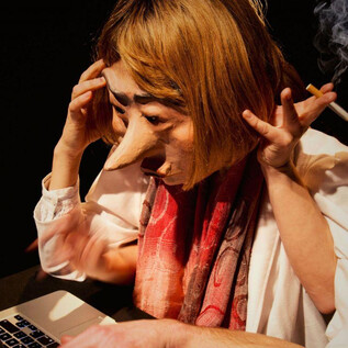 A person with a large mask is sitting in front of a laptop. They look stressed and are holding a cigarette in their hand. | © Debra Tchotchov