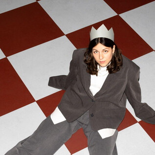 A person is sitting on a chessboard floor wearing a crown hat. They are dressed in a gray suit and looking directly at the camera. | © Sima Prodinger