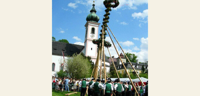 A traditional celebration with people in traditional costumes erecting a Maypole. In the background is a church under a cloudy sky. | © TMK_Aigen