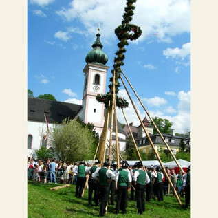 A traditional celebration with people in traditional costumes erecting a Maypole. In the background is a church under a cloudy sky. | © TMK_Aigen