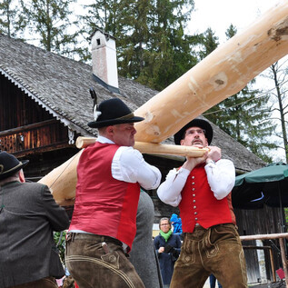 Men in traditional costumes are lifting a heavy wooden beam. In the background, a rustic house and nature can be seen. | © Salzburger Freilichtmuseum