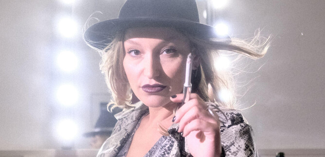 A woman with a hat and a confident expression stands in front of a lit mirror. She holds a pen in her hand and has a fashionable look. | © Christopher Glanzl