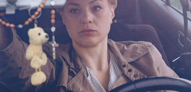 A woman is sitting at the wheel of a car and looking thoughtfully. In the background, a car seat and a hanging keychain can be seen. | © Apollonia Theresa Bitzan