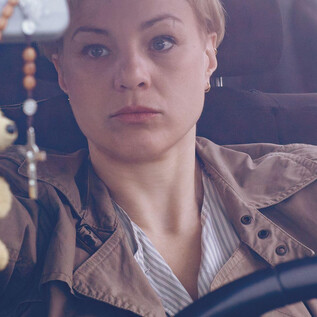 A woman is sitting at the wheel of a car and looking thoughtfully. In the background, a car seat and a hanging keychain can be seen. | © Apollonia Theresa Bitzan