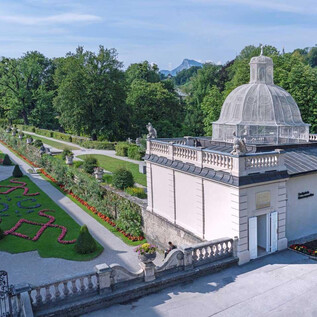 A beautiful garden with carefully arranged flower beds and lots of greenery. In the background, a classic building can be seen, surrounded by trees and plants. | © Stadt Salzburg