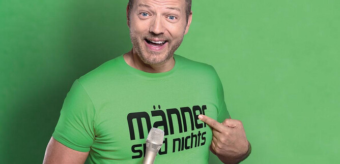 A smiling man is wearing a green T-shirt with the slogan "Men are nothing." He is holding a microphone and pointing at it with his finger. | © S-Promotion Event GmbH