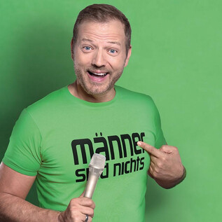 A smiling man is wearing a green T-shirt with the slogan "Men are nothing." He is holding a microphone and pointing at it with his finger. | © S-Promotion Event GmbH