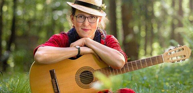 A woman is sitting in the grass holding a guitar. She is wearing a hat and a red blouse, surrounded by trees and flowers. | © Gregor Wiebe