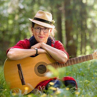 A woman is sitting in the grass holding a guitar. She is wearing a hat and a red blouse, surrounded by trees and flowers. | © Gregor Wiebe