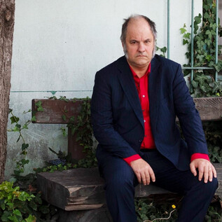A man is sitting on a wooden bench under a tree. He is wearing a dark suit and a red shirt while thoughtfully looking at the camera. | © Stephan Minx
