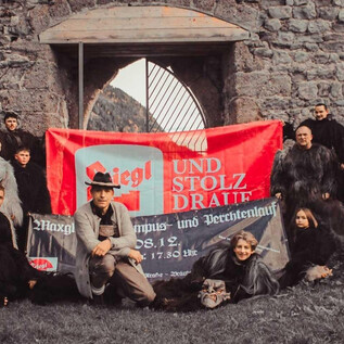 A group of people is standing in front of an old castle and holding a large banner. Everyone is wearing striking, dark clothing and posing for the photo.