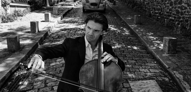 A cellist is playing on a cobblestone street in a picturesque town. In the background, an older car and characteristic buildings can be seen. | © Anoush Abrar