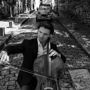 Ein Cellist spielt auf einer gepflasterten Straße in einer malerischen Stadt. Im Hintergrund ist ein älteres Auto und charakteristische Gebäude zu sehen. | © Anoush Abrar