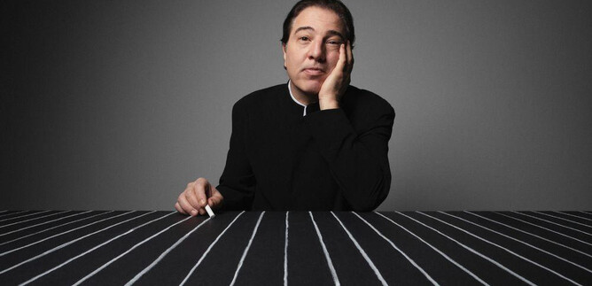 A pensive man sits at a table with black and white stripes. He has one hand on his face and looks serious. | © Fethi Karaduman