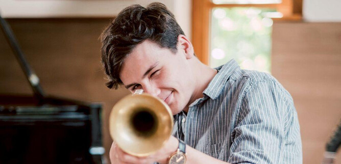 A young man is enthusiastically playing a trumpet. In the background, a piano and a warm, friendly room atmosphere can be seen. | © Kunsthilfe Salzburg