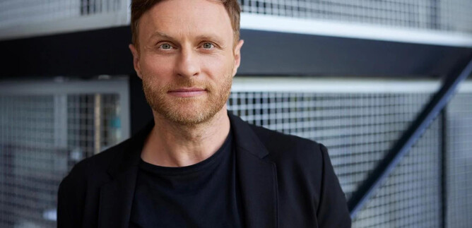 A man with a short beard is standing in front of a modern, textured wall. He is wearing a black blazer and smiling slightly. | © Steffen Jänicke