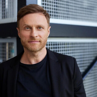 Egy rövid szakállú férfi egy modern, strukturált fal előtt áll. Fekete zakót visel és enyhén mosolyog. | © Steffen Jänicke