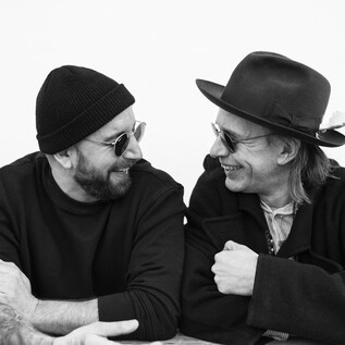 Two men are laughing and looking at each other friendly. They are wearing sunglasses and stylish hats. | © Daniela Matejschek