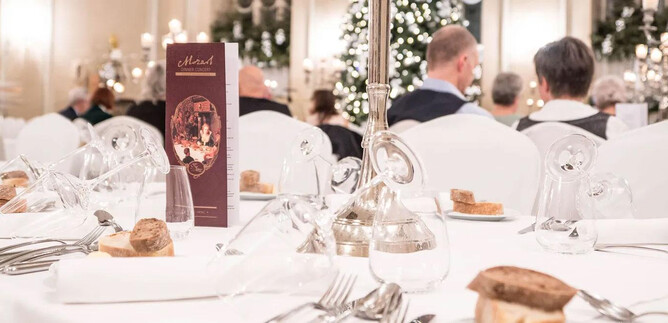 A festively set table in an elegant restaurant. In the background, guests can be seen attending a festive dinner. | © St. Peter Stiftskulinarium