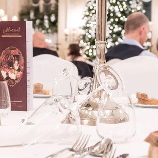 A festively set table in an elegant restaurant. In the background, guests can be seen attending a festive dinner. | © St. Peter Stiftskulinarium