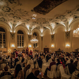 An elegant banquet hall with many guests sitting at festively decorated tables. The ceiling is artistically embellished and in the background, a band can be seen. | © Michael Groessinger