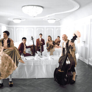 A group of people in elegant clothing stands in a festive room. Some are holding musical instruments, while others pose and appear cheerful. | © Nancy_Horowitz