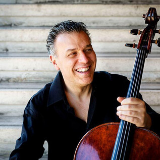 A smiling cellist sits on a staircase holding his cello. He wears a black shirt and radiates joy. | © Musacchio,-Ianniello-e-Pasqualini