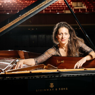 A talented pianist sits at an open grand piano in a concert hall. Her gaze is focused and she radiates enthusiasm for the music. | © Guillaum Perret