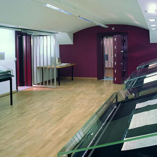 A modern exhibition space with wooden flooring and red walls. Glass cases showcase various documents and exhibits. | © ISM