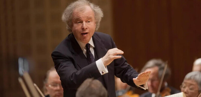 A conductor leads an orchestra with passionate gestures. In the background, musicians with their instruments can be seen. | © Priska Ketterer