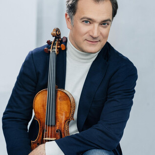 A musician is sitting with a violin in hand. He is wearing an elegant blazer and looking kindly at the camera. | © Universal Music