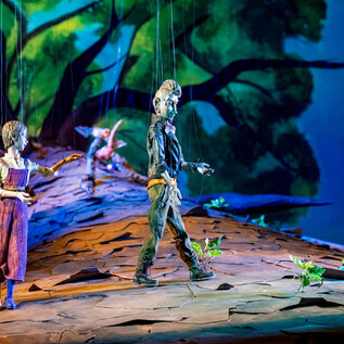A stage with two puppets standing on a large tree trunk. In the background, there is a vibrant forest scene with colorful hues. | © Bernhard Mueller