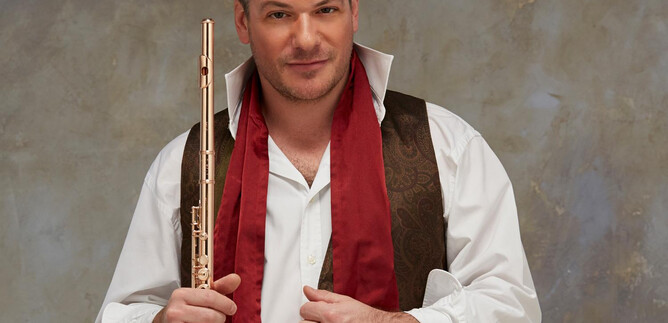 A man holds a flute in his hand and wears an elegant outfit with a red scarf. The background is neutral and slightly textured. | © Fabien Monthubert