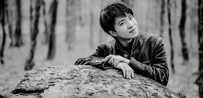 A young man leans against a tree trunk in the forest. The scene is in black and white and conveys a calm atmosphere. | © Marco Borggreve