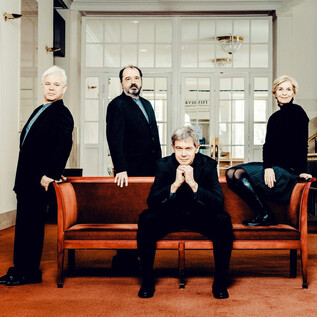 A group of four people is standing and sitting on a sofa in a bright interior. They are all wearing black clothing and looking at the camera. | © Andrej Grilc