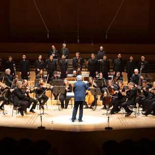 An orchestra and a choir perform on a stage. The conductor leads the musicians and singers in formal attire. | © Penar Royal