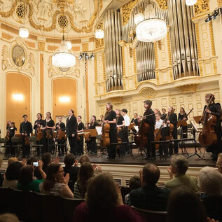 An orchestra on the stage of an elegant concert hall. The audience sits attentively and applauds. | © Wol
