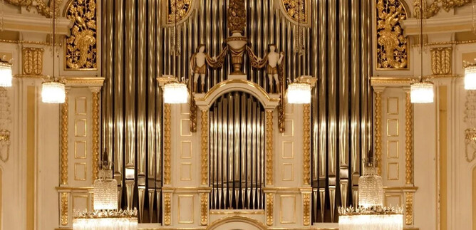 An impressive organ with golden decorations in a festive room. The walls are brightly and elegantly designed, featuring hanging lights. | © Wolfgang Lienbacher