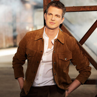 A well-dressed man stands casually against a modern background. He is wearing a brown suede jacket and a white shirt. | © Simon Fowler
