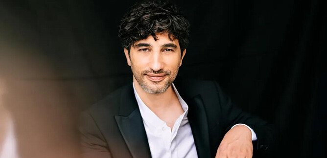 A man in a black suit is sitting in front of a dark background. He is smiling warmly and has curly hair. | © Christoph Köstlin